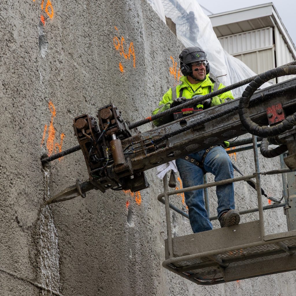 A drill operator operates drilling equipment from a work platform to install hollow bar anchors into a reinforced concrete wall.