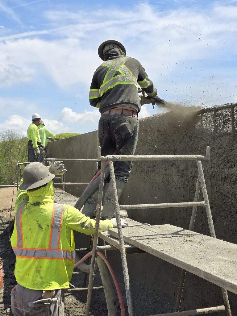 A nozzleman applying shotcrete to a retaining wall from an elevated platform.