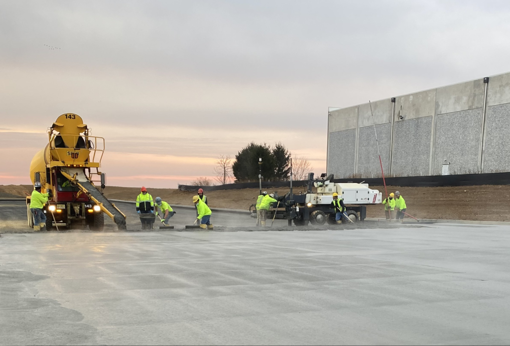 Multiple workers raking a large concrete slab actively being poured.