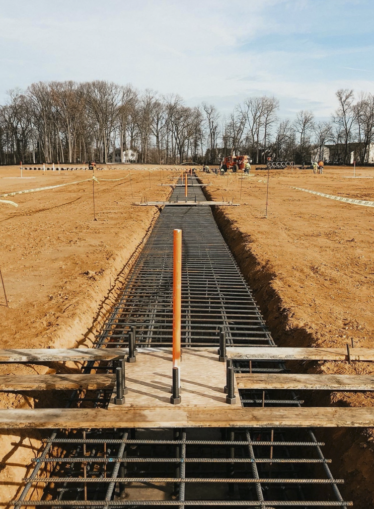 Reinforcing steel and an orange utility conduit are installed within a long foundation trench before concrete placement.