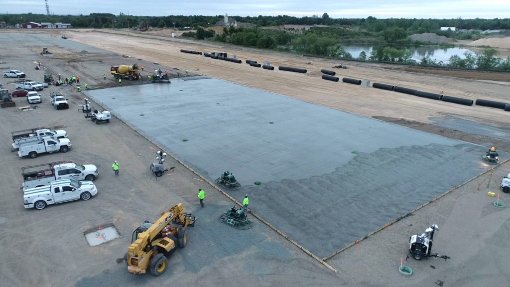 Half poured concrete slab featured ride-on trowel finishers, multiple work trucks, and concrete mixer trucks.